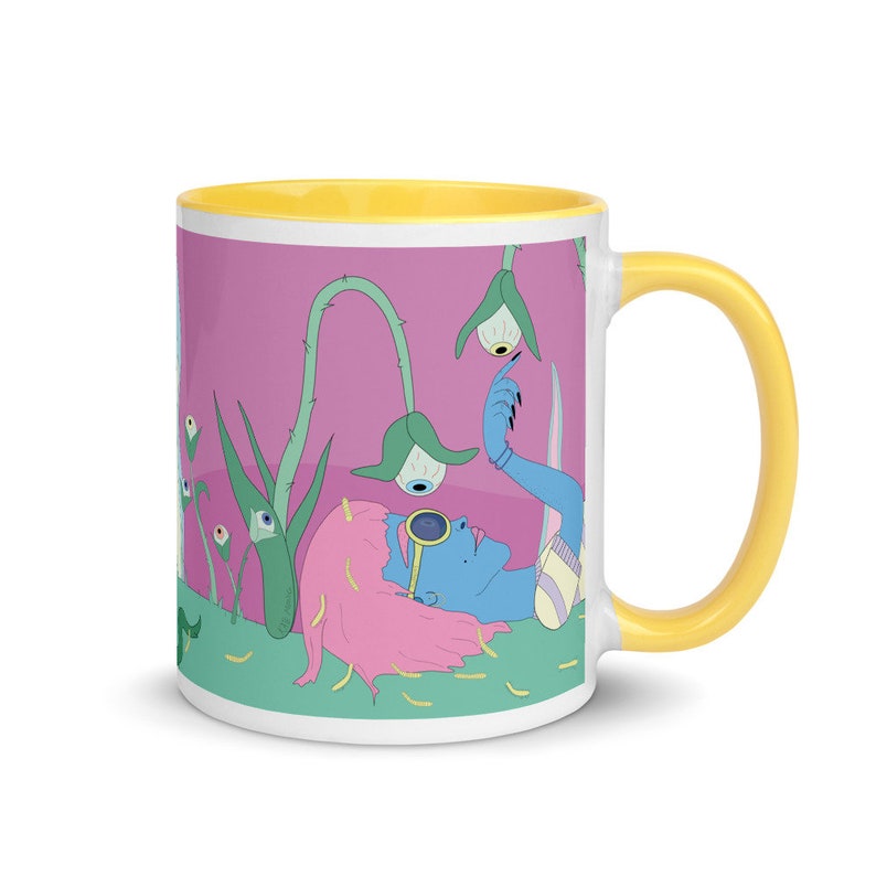 May include: A white ceramic mug with a yellow rim and handle. The mug features a colorful illustration of a person with pink hair lying in a field of green plants with eyes. The background is pink.