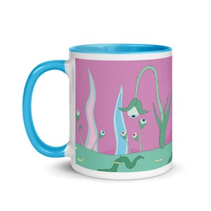 May include: A white ceramic mug with a light blue rim and handle. The mug features a pink background with a whimsical illustration of green plants with blue and white eyes.