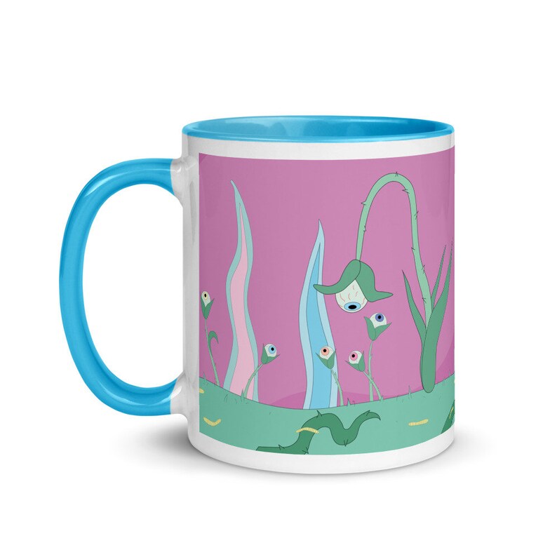 May include: A white ceramic mug with a light blue rim and handle. The mug features a pink background with a whimsical illustration of green plants with blue and white eyes.
