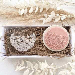 Pode incluir: Dois toppers redondos para bolos, de cor branca, com motivos florais, numa caixa branca com enchimento de papel castanho. Um topper tem a forma de um coração e o outro é um círculo.