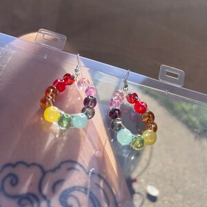 Puede incluir: Un par de pendientes de cuentas de colores. Cada pendiente presenta una forma de lágrima hecha de cuentas de varios colores, incluyendo rojo, amarillo, verde y azul. Los pendientes tienen ganchos plateados.