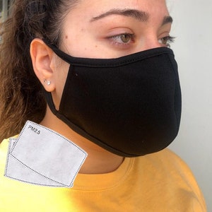 Pode incluir: Uma máscara facial preta com um filtro PM2.5 branco. A máscara é feita de um tecido macio e respirável e tem laços para as orelhas ajustáveis.