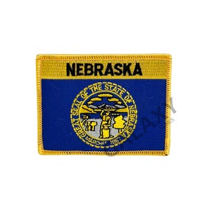 May include: A blue and gold embroidered patch featuring the state seal of Nebraska and the word "NEBRASKA" in black lettering.