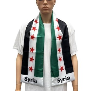 May include: A person wearing a white shirt with a black and green scarf. The scarf has the Syrian flag on it with red stars and the word "Syria" written on it.