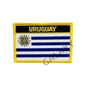 Pode incluir: Um remendo bordado amarelo com a palavra "URUGUAY" em letras pretas sobre a bandeira uruguaia. A bandeira é branca com um sol azul no canto superior esquerdo e nove faixas azuis horizontais.