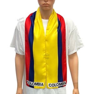 May include: A yellow, blue, and red striped scarf with the word "COLOMBIA" printed on each end. The scarf is worn by a person wearing a white t-shirt.