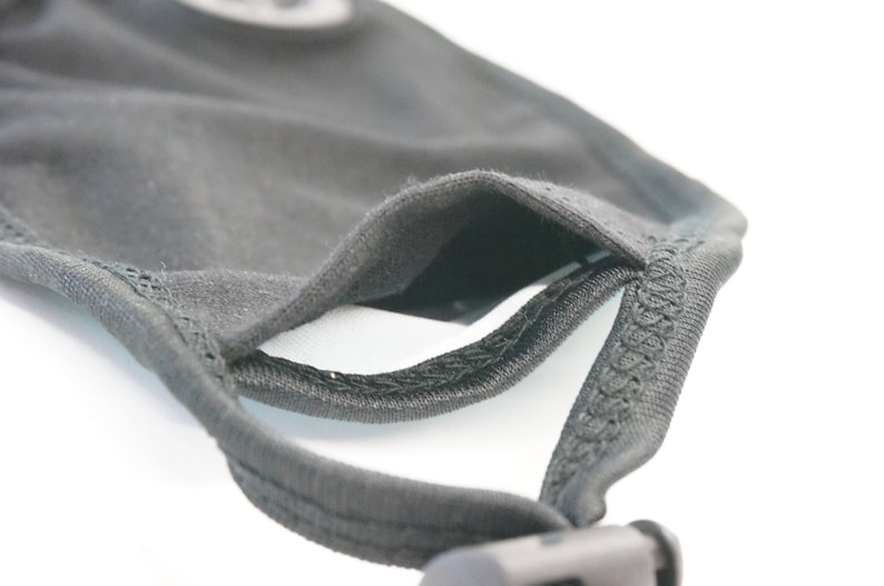 May include: Close-up of a black face mask with a black elastic strap and a black adjustable buckle. The mask has a double layer of fabric and a pocket for a filter.