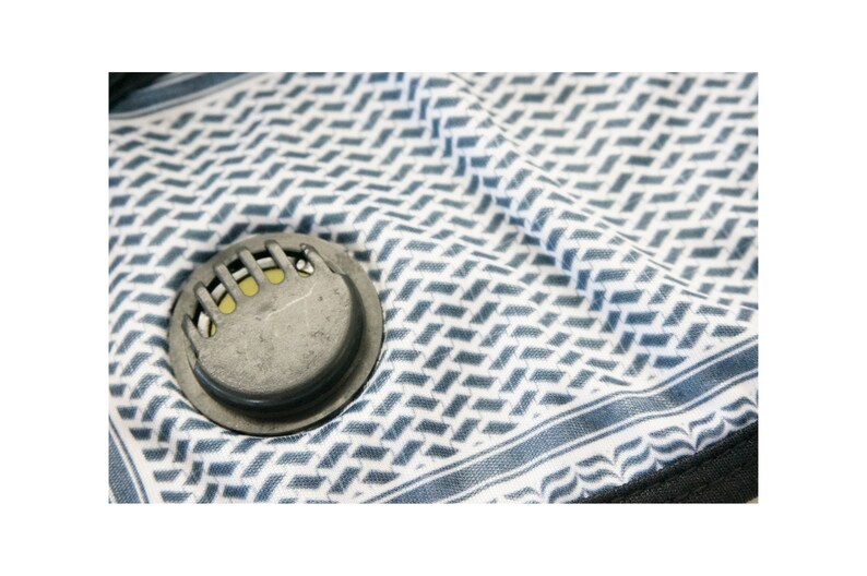 May include: A close-up of a gray and white patterned face mask with a round, gray, metal valve. The valve has a series of small, yellow, rectangular openings.