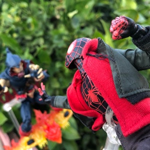 May include: A red and black action figure of Spider-Man wearing a red jacket and black pants, with a black spider logo on the chest. The figure is posed in a fighting stance with its right fist raised. The figure is standing on a clear plastic stand. In the background, a blue and red action figure of a villain is standing on a fiery base.