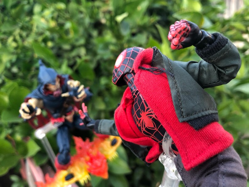 May include: A red and black action figure of Spider-Man wearing a red jacket and black pants, with a black spider logo on the chest. The figure is posed in a fighting stance with its right fist raised. The figure is standing on a clear plastic stand. In the background, a blue and red action figure of a villain is standing on a fiery base.