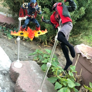 May include: Two action figures, one in a red and black suit with a red mask, and the other in a blue and black suit with a red mask and a yellow and orange flame design, are posed in a fighting stance. The figures are on clear plastic stands.