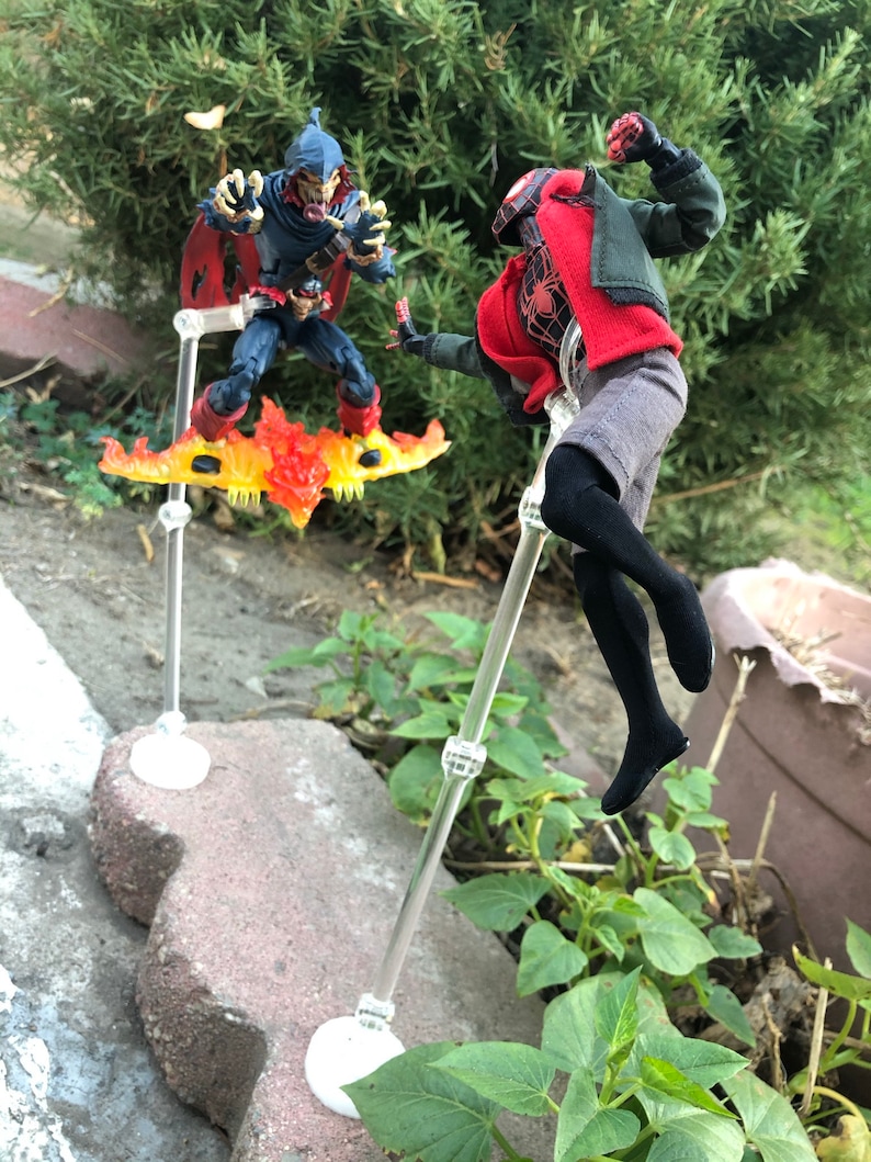 May include: Two action figures, one in a red and black suit with a red mask, and the other in a blue and black suit with a red mask and a yellow and orange flame design, are posed in a fighting stance. The figures are on clear plastic stands.