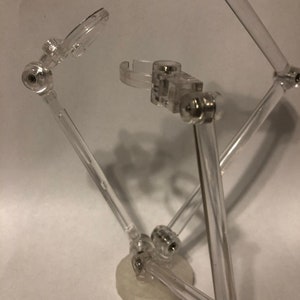 May include: A clear plastic action figure stand with adjustable arms and a circular base. The stand is designed to hold and pose action figures.