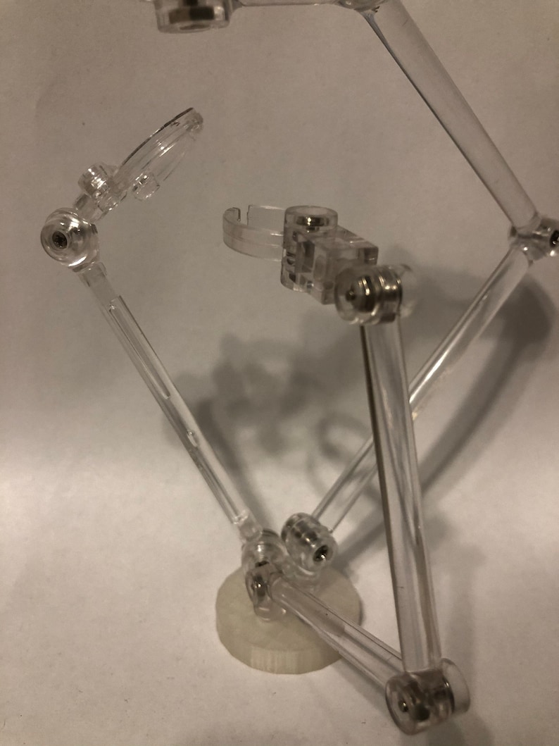 May include: A clear plastic action figure stand with adjustable arms and a circular base. The stand is designed to hold and pose action figures.