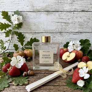 May include: A square glass reed diffuser bottle with a gold cap and a label reading WICK & BAR INDIAN SUMMER 7 OZ DIFFUSER. The bottle is accompanied by a bundle of white reed sticks tied with a gold ribbon, surrounded by fresh strawberries, sliced apples, white flowers, and oak leaves.