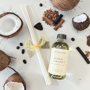 May include: A flat lay featuring a Wick & Bar Santal + Coconut diffuser with reeds, a yellow ribbon, and coconut halves. The bottle is 4 oz. and includes sandalwood, cedar, and teak notes.
