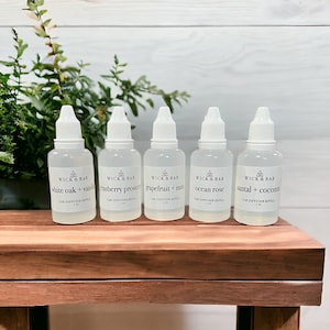 May include: Five small clear glass bottles with white caps and labels. The labels read "Wick & Bar" and list different scents: "white oak + vanilla", "cranberry prosecco", "grapefruit + mint", "ocean rose", and "santal + coconut".