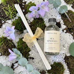 May include: A clear glass bottle labeled "SEA MINERALS" with a black cap, next to a bundle of white diffuser sticks tied with a gold ribbon. The image includes purple flowers, green moss, and white granules, creating a natural aesthetic.