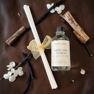 May include: A clear glass diffuser bottle with a black cap, labeled "WHITE OAK + VANILLA." White reeds are tied with a gold ribbon. The image includes decorative elements like wood pieces, vanilla beans, and white flowers, all on a brown surface.