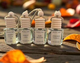 May include: Four small glass car diffusers with wooden tops and labels. The labels read "Brown Sugar + Fig", "Forest Chai", "Dark Orchard", and "Ambered Vanilla". Each diffuser has a string for hanging.