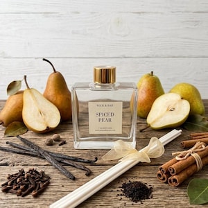 May include: A clear glass reed diffuser bottle with a gold cap and a label reading WICK & BAR SPICED PEAR 7 OZ DIFFUSER. The bottle is surrounded by whole and halved pears, vanilla beans, cloves, cinnamon sticks, and white diffuser reeds.
