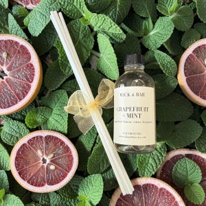 May include: Aromatherapy diffuser set with a bottle labeled "WICK & BAR GRAPEFRUIT + MINT" and reed sticks tied with a gold ribbon. The image includes sliced grapefruits and green mint leaves, creating a fresh, natural aesthetic.