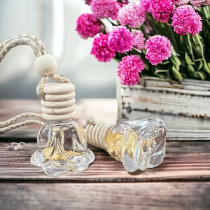 May include: Two clear glass car air fresheners shaped like roses, filled with gold flakes, hanging from a white string. The air fresheners are on a wooden surface with pink flowers in the background.