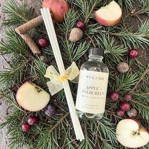 May include: A clear glass diffuser bottle with a black cap, labeled "Apple + Evergreen." The bottle is surrounded by pine needles, apple slices, cranberries, and cinnamon sticks. A bundle of white diffuser sticks is tied with a gold ribbon.