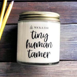 May include: A white candle jar with a gold lid and a black label that reads "tiny human tamer" and a triangle logo with the text "Wick & Bar".