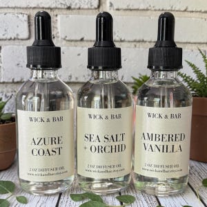 May include: Three clear glass diffuser oil bottles with black droppers. Each bottle has a label with the text "WICK & BAR" and a scent name: "AZURE COAST", "SEA SALT + ORCHID", and "AMBERED VANILLA". The bottles are 2 oz.