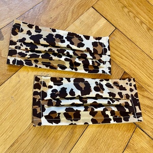 May include: Two brown and white leopard print face masks with white stitching. The masks are pleated and have a rectangular shape.