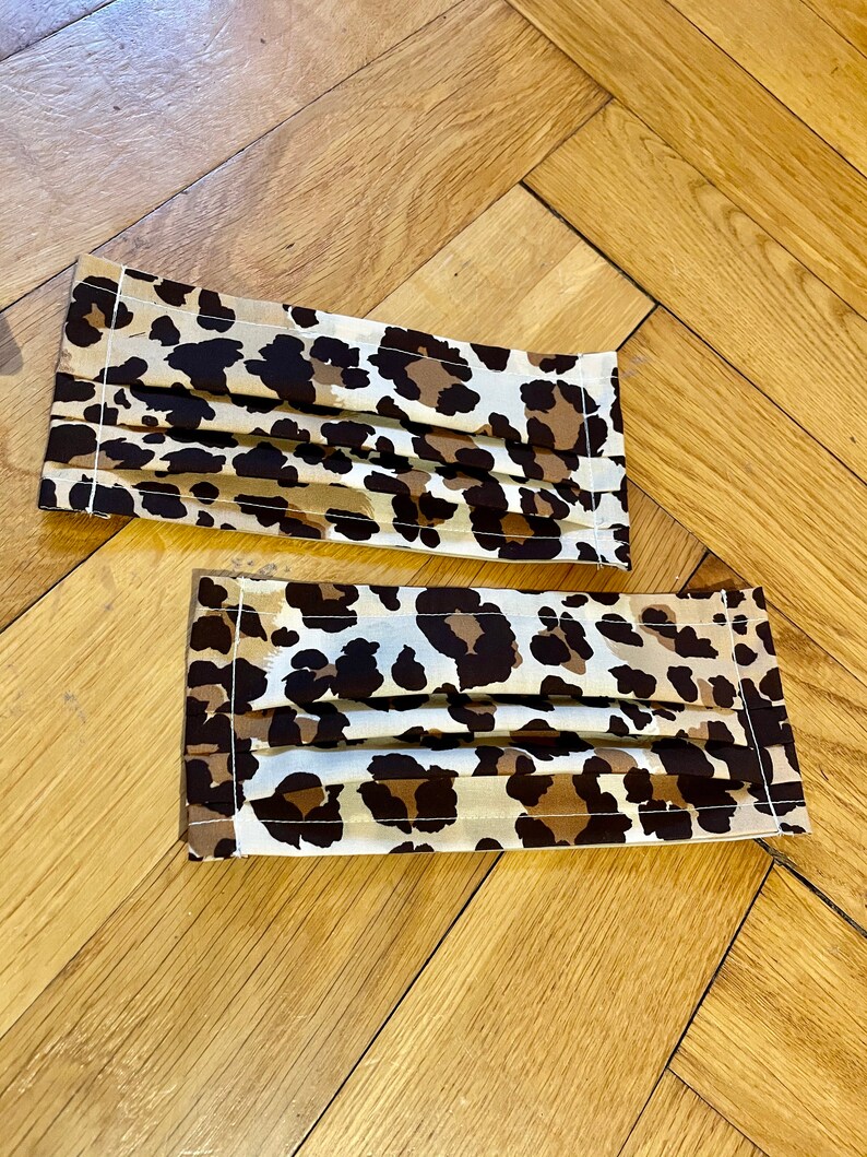 May include: Two brown and white leopard print face masks with white stitching. The masks are pleated and have a rectangular shape.