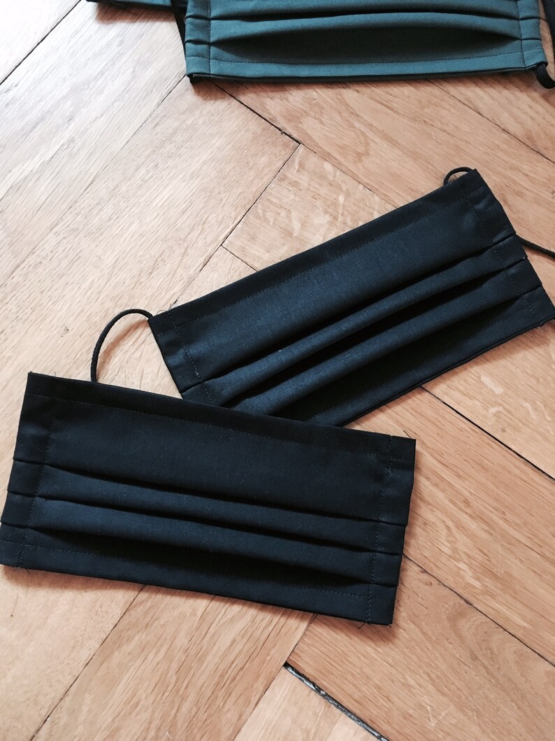 May include: Two pleated fabric face masks, one black and one dark green, laid out on a wooden surface.