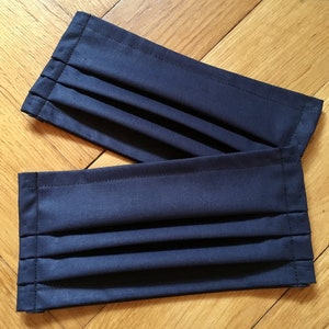 May include: Two dark blue fabric face masks with pleats. The masks are folded and lying on a wooden surface.