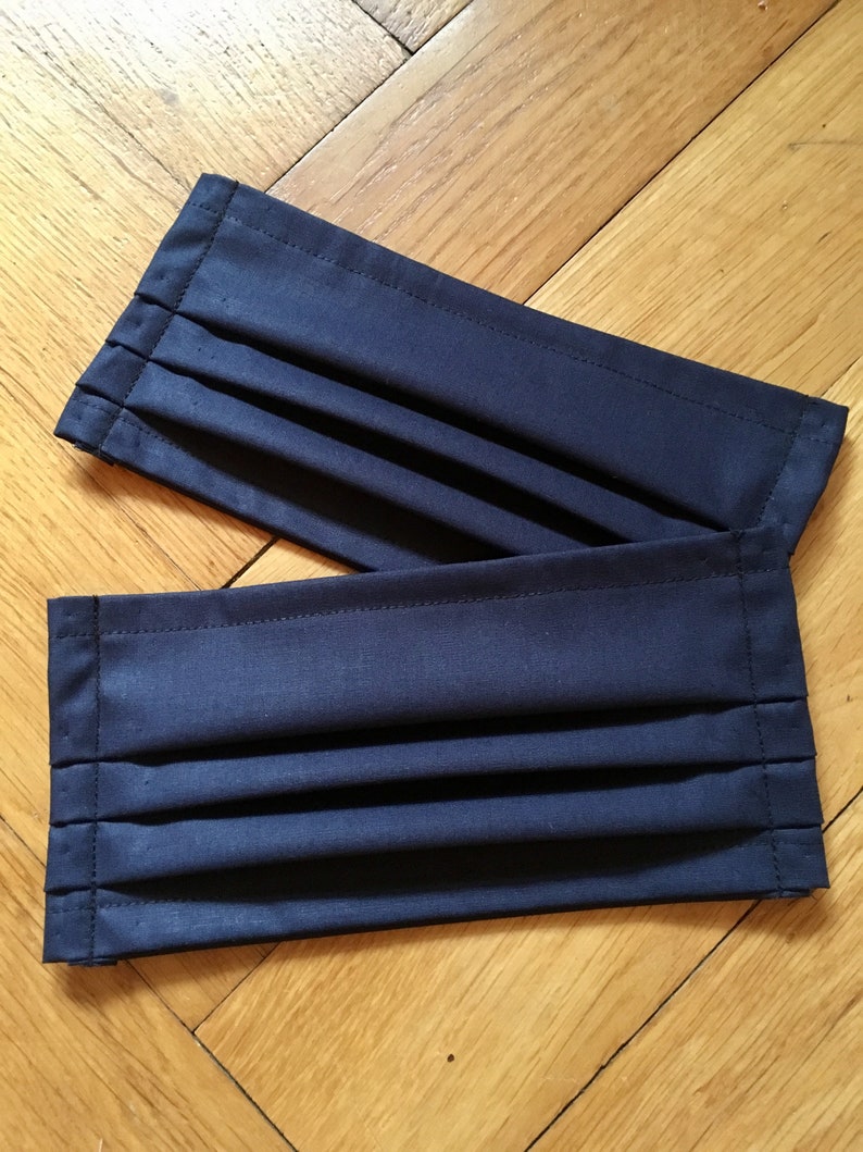 May include: Two dark blue fabric face masks with pleats. The masks are folded and lying on a wooden surface.