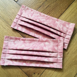 May include: Two pink face masks with a tie-dye pattern. The masks are pleated and have a white background with pink swirls.