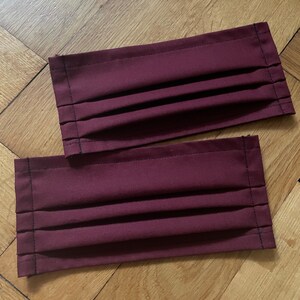 May include: Two burgundy fabric face masks with pleats and black stitching.