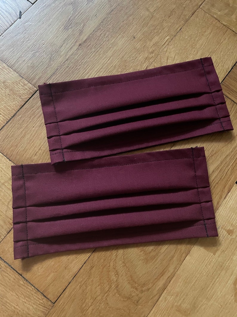 May include: Two burgundy fabric face masks with pleats and black stitching.