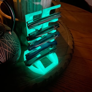 May include: A glowing green plastic stand with multiple slots for holding pocket knives. The stand is on a wooden surface.