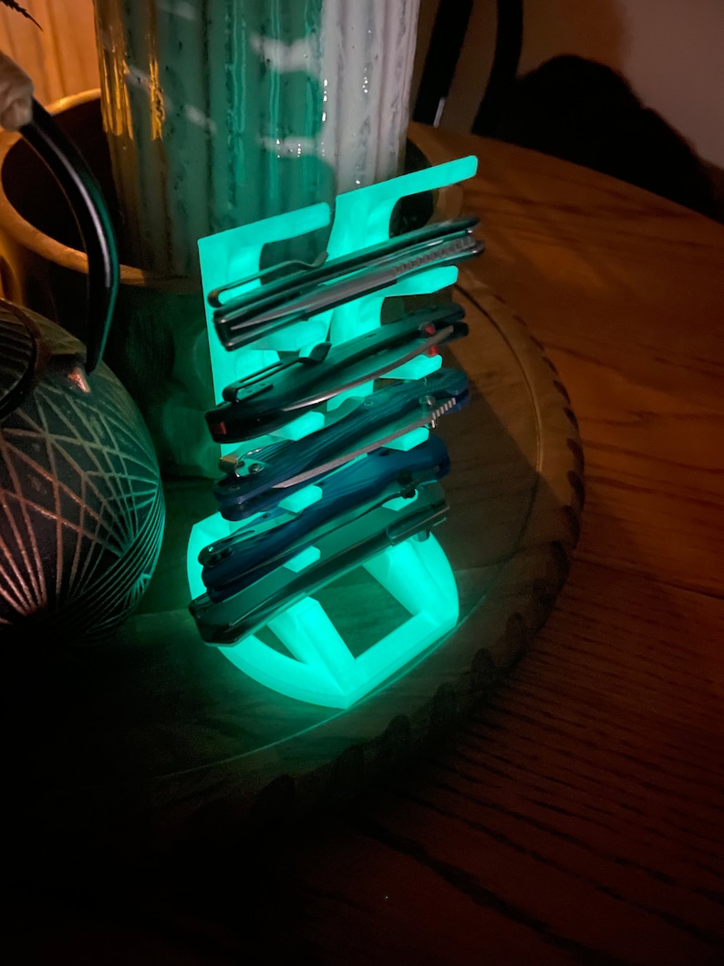 May include: A glowing green plastic stand with multiple slots for holding pocket knives. The stand is on a wooden surface.