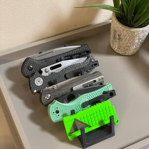 May include: A black plastic knife holder with five knives. The knives are black, gray, and green. The holder is on a gray surface.