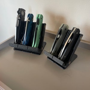 May include: Two black knife stands with a textured surface, each holding multiple pocket knives. The knives are in various colors, including black, green, and blue.