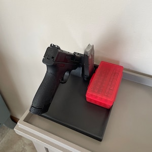 May include: A black handgun with a textured grip rests on a black tray, next to a magazine and a red ammunition case. The items are on a light gray table against a white wall. The handgun is a semi-automatic pistol.