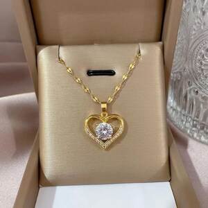 Puede incluir: Un collar dorado con un colgante en forma de corazón. El colgante presenta una gran gema transparente en el centro, rodeada de piedras más pequeñas y transparentes. El collar se presenta en una caja de joyería beige.