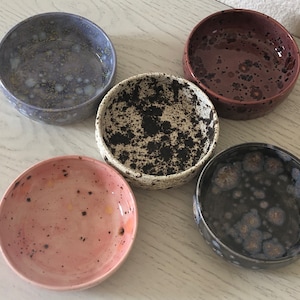 May include: Five small ceramic bowls with speckled patterns. The bowls are in shades of blue, brown, white, black, and pink.