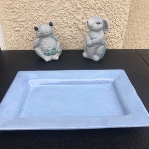 May include: A light blue ceramic rectangular serving tray with a raised rim. The tray is sitting on a black surface. There is a gray frog figurine and a gray rabbit figurine on the surface behind the tray.