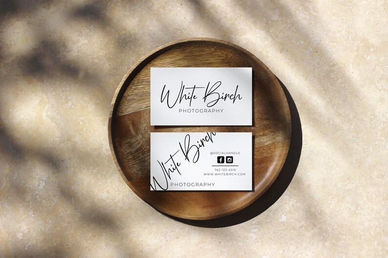 K&ouml;nnte beinhalten: Zwei wei&szlig;e Visitenkarten mit schwarzem Text auf einem Holzteller. Die obere Karte lautet "White Birch Photography". Die untere Karte lautet "White Birch Photography" mit einem Social-Media-Handle, einer Telefonnummer und einer Website-Adresse.