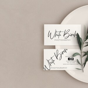 K&ouml;nnte beinhalten: Zwei wei&szlig;e Visitenkarten mit dem Text "White Birch Photography" in schwarzer Kursivschrift. Die obere Karte tr&auml;gt den Text "Photography" unter dem Namen. Die untere Karte enth&auml;lt Social-Media-Symbole und Kontaktinformationen. Die Karten liegen auf einem wei&szlig;en Teller mit einem gr&uuml;nen Olivenzweig.