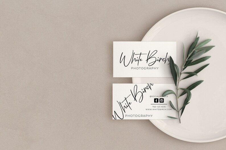 K&ouml;nnte beinhalten: Zwei wei&szlig;e Visitenkarten mit dem Text "White Birch Photography" in schwarzer Kursivschrift. Die obere Karte tr&auml;gt den Text "Photography" unter dem Namen. Die untere Karte enth&auml;lt Social-Media-Symbole und Kontaktinformationen. Die Karten liegen auf einem wei&szlig;en Teller mit einem gr&uuml;nen Olivenzweig.