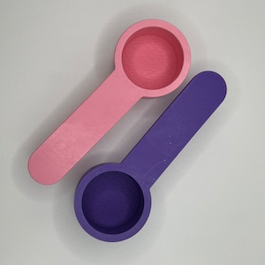 Peut inclure: Deux cuill&egrave;res doseuses avec des bols ronds et des manches plats. Une cuill&egrave;re est rose clair et l'autre est violet fonc&eacute;. Les deux articles pr&eacute;sentent une finition de surface textur&eacute;e typique des objets imprim&eacute;s en 3D, avec un bol circulaire.
